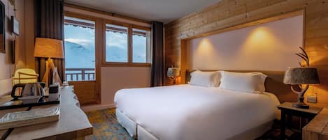 A hotel room with a large bed, wooden walls, a window with a view of the mountains, a desk with a lamp, and a patterned rug.