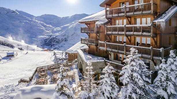Eine Holzchalet mit mehreren Balkonen, umgeben von schneebedeckten Bergen und einer Skipiste.