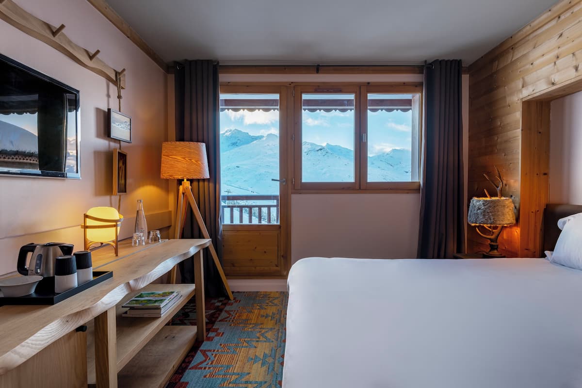 A hotel room with a bed, a wooden desk with a lamp, a view of snow-covered mountains, and a window with curtains.