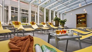 A sunlit indoor seating area with yellow chairs and tables, a glass ceiling, and a wooden bookshelf. - Dorint Seehotel Binz-Therme Binz/Rügen (Binz)