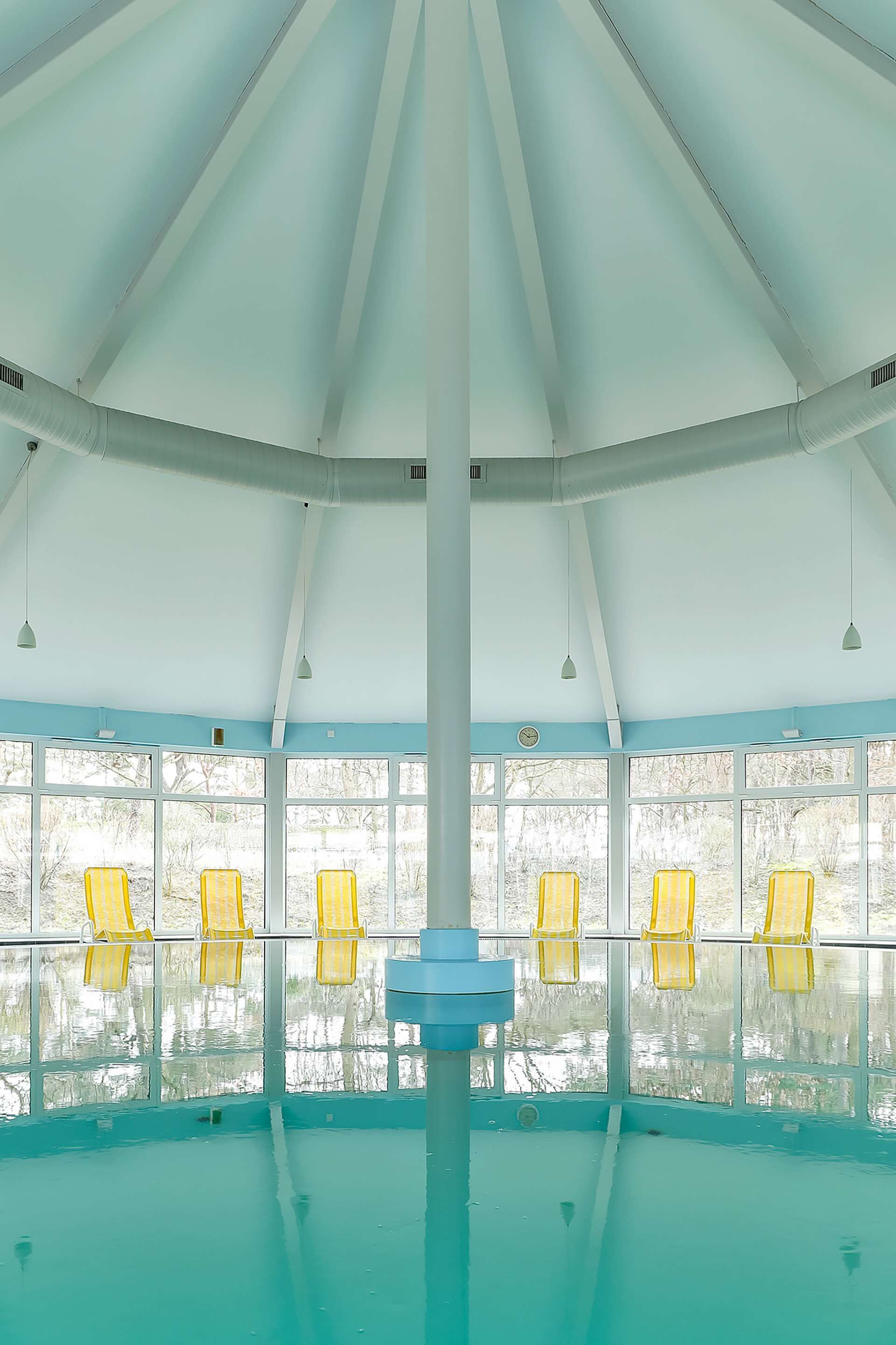 En indendørs swimmingpool med højt til loftet, store vinduer og gule stole langs bassinets kant.