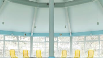 An indoor swimming pool with a high ceiling, large windows, and yellow chairs along the poolside.