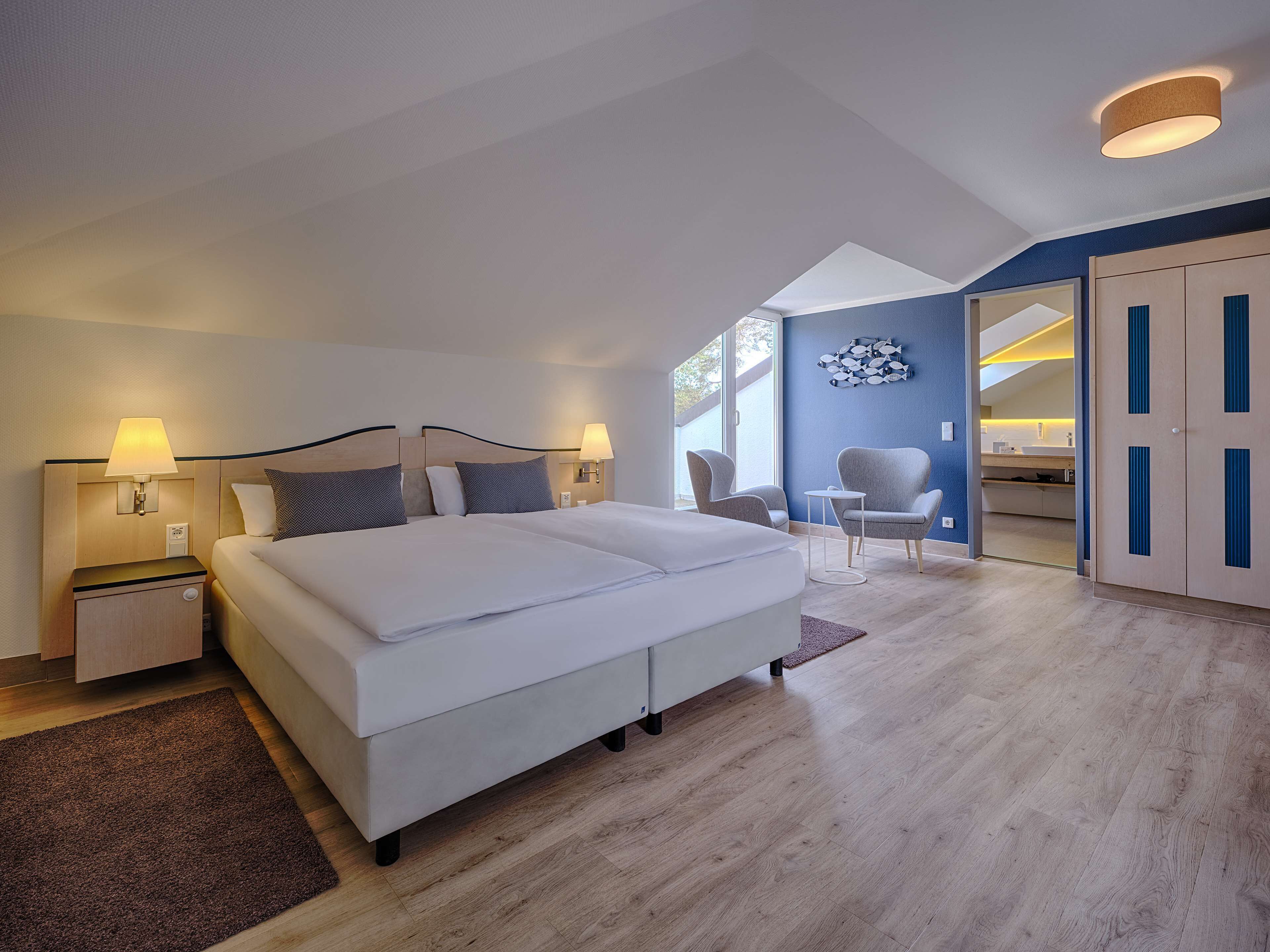 Una habitación de hotel moderna con una cama grande, dos mesitas de noche con lámparas, un área de estar con sillas y vistas al exterior.