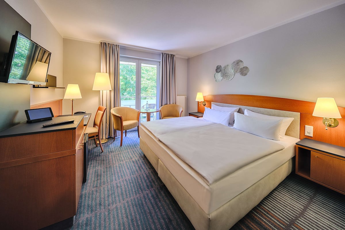 Une chambre d’hôtel avec un grand lit, un bureau avec un ordinateur portable, deux chaises, une télévision et une fenêtre avec des rideaux.