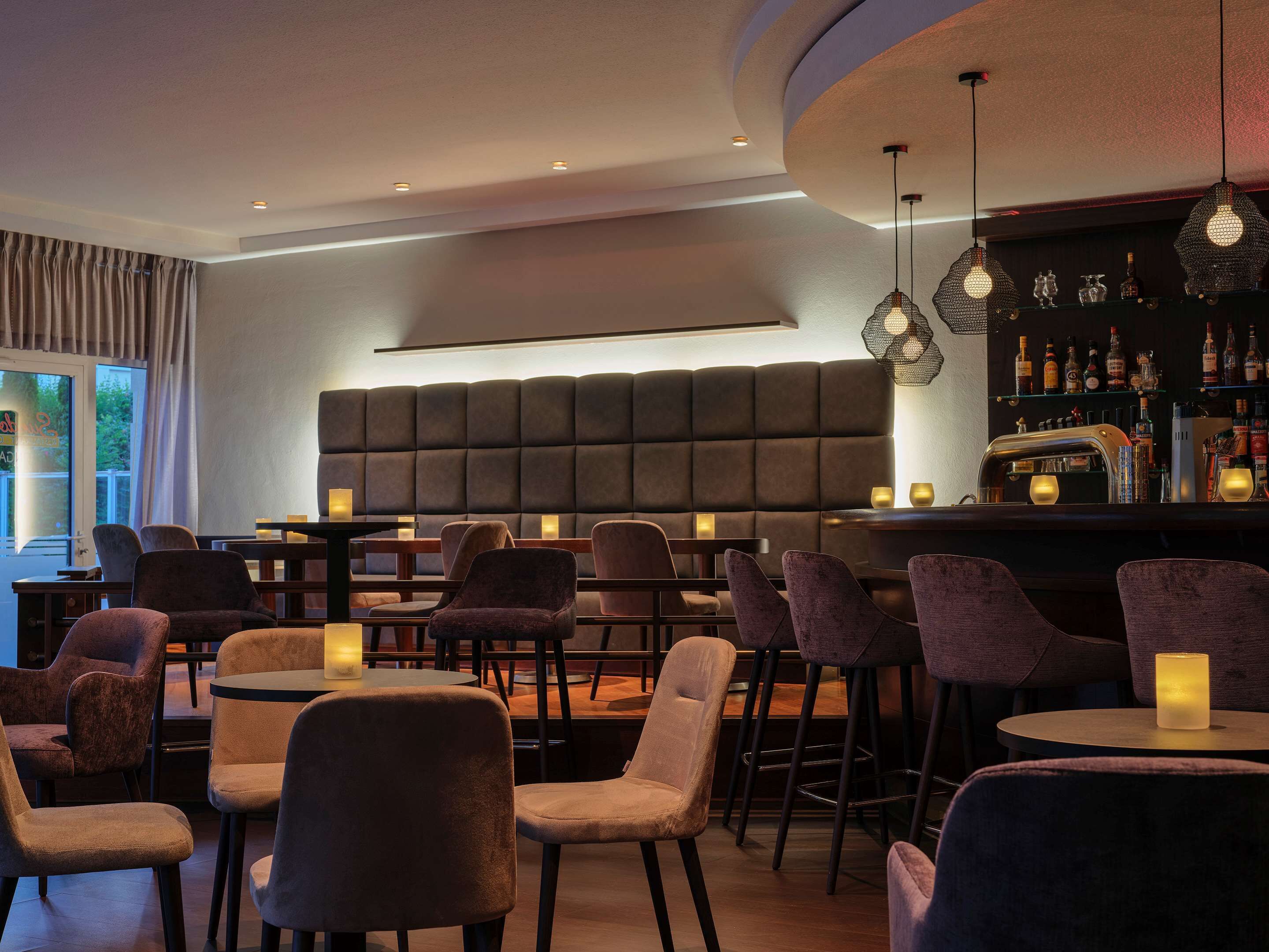 A modern bar area with cushioned seating, pendant lighting, and a bar stocked with bottles.