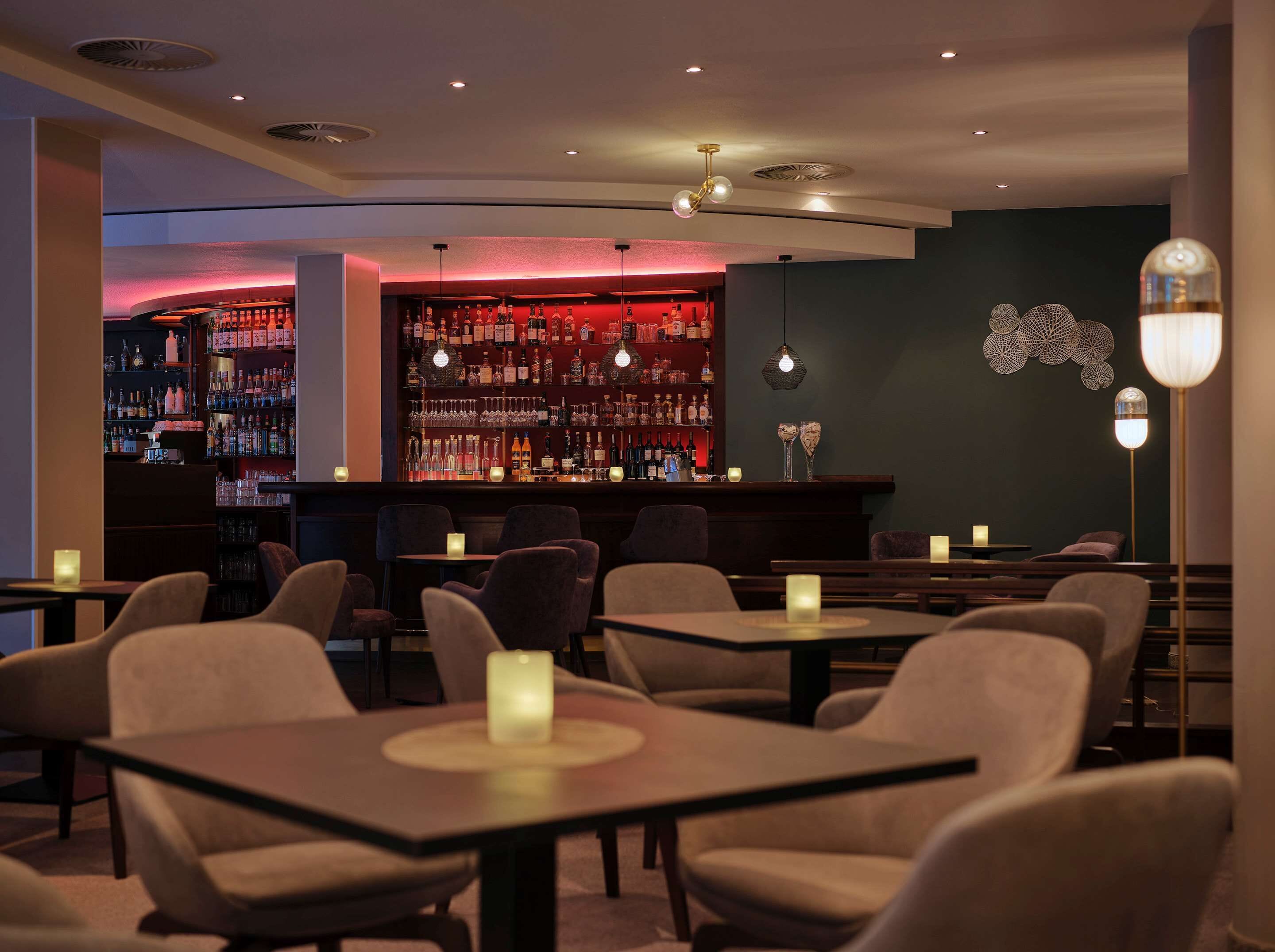 A bar area with a dark wall and shelves of bottles, tables with chairs, and a decorative light fixture.