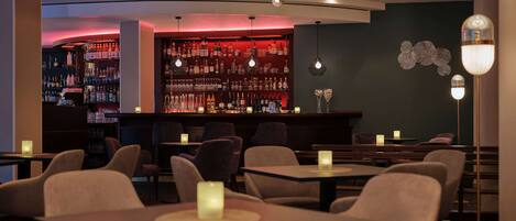 A bar area with a dark wall and shelves of bottles, tables with chairs, and a decorative light fixture.