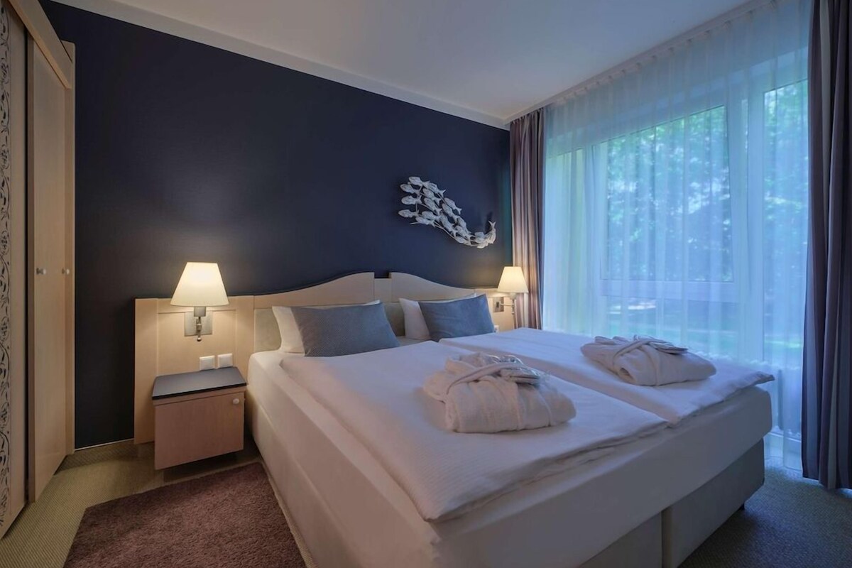 A hotel room with a large bed, two bedside tables, a wall-mounted lamp, and a window with curtains.