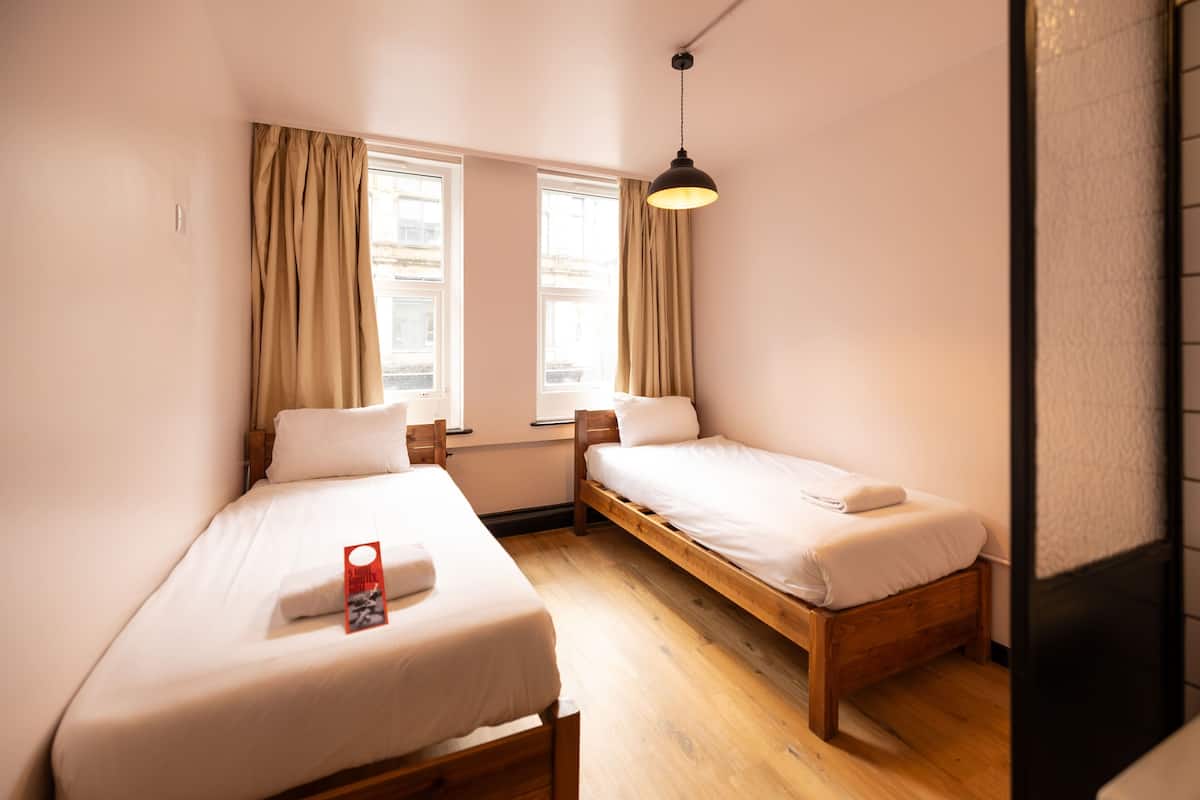 A small, clean room with two single beds, a wooden floor, and a window with curtains.