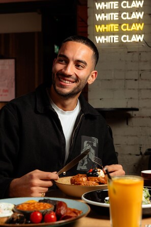 Une personne est assise à une table devant une assiette de nourriture, un verre de jus d’orange et une fourchette.