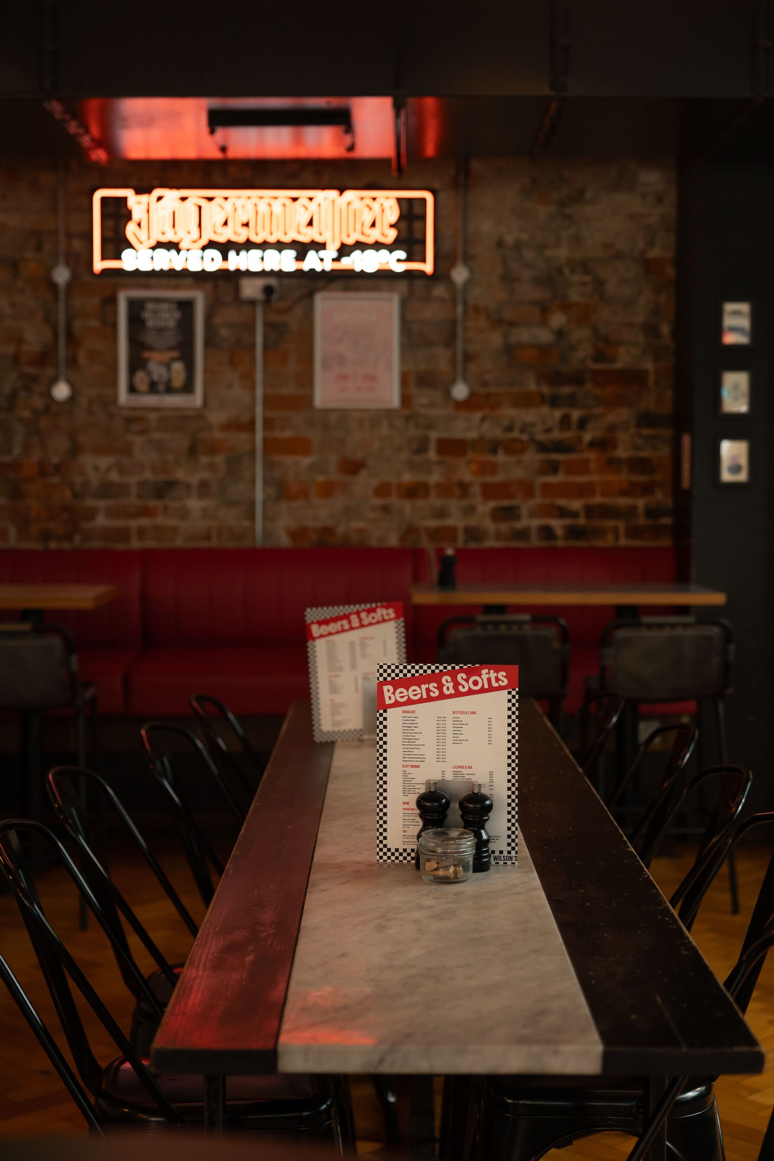 A restaurant with a long wooden table, black chairs, and a menu stand displaying 'Beers & Softs'.