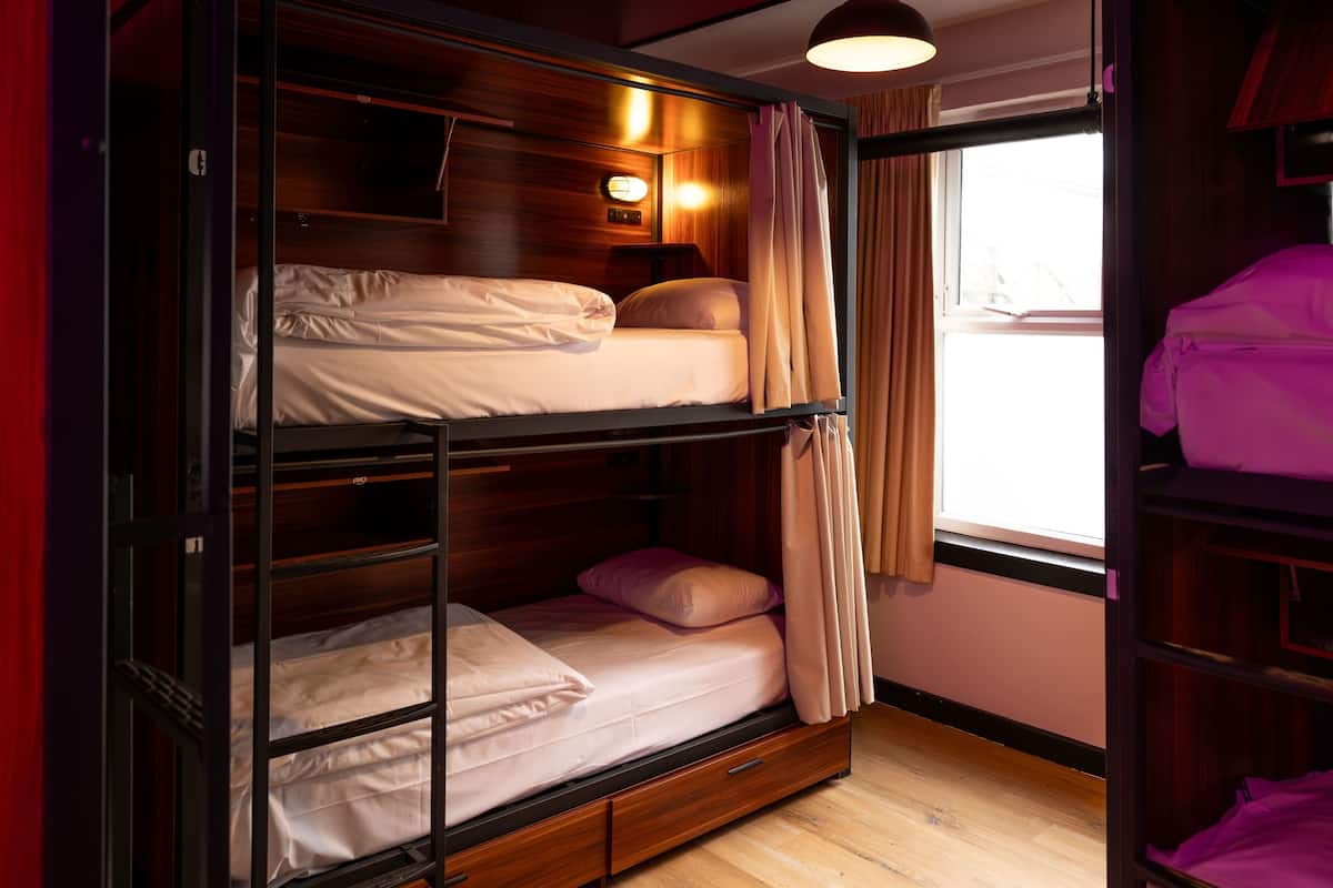 A bunk bed room with wooden headboard, a window with curtains, and a bedside lamp.