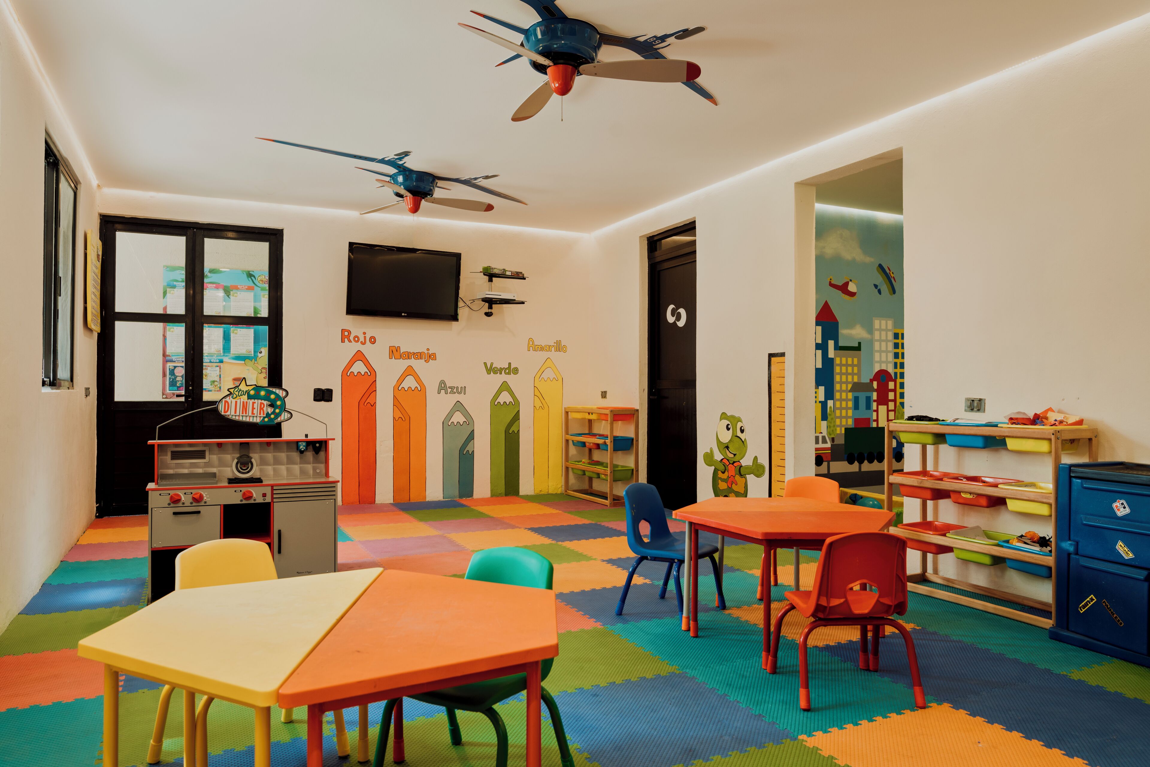 A children's playroom with colorful mats, tables, chairs, ceiling fans, and educational wall decorations.