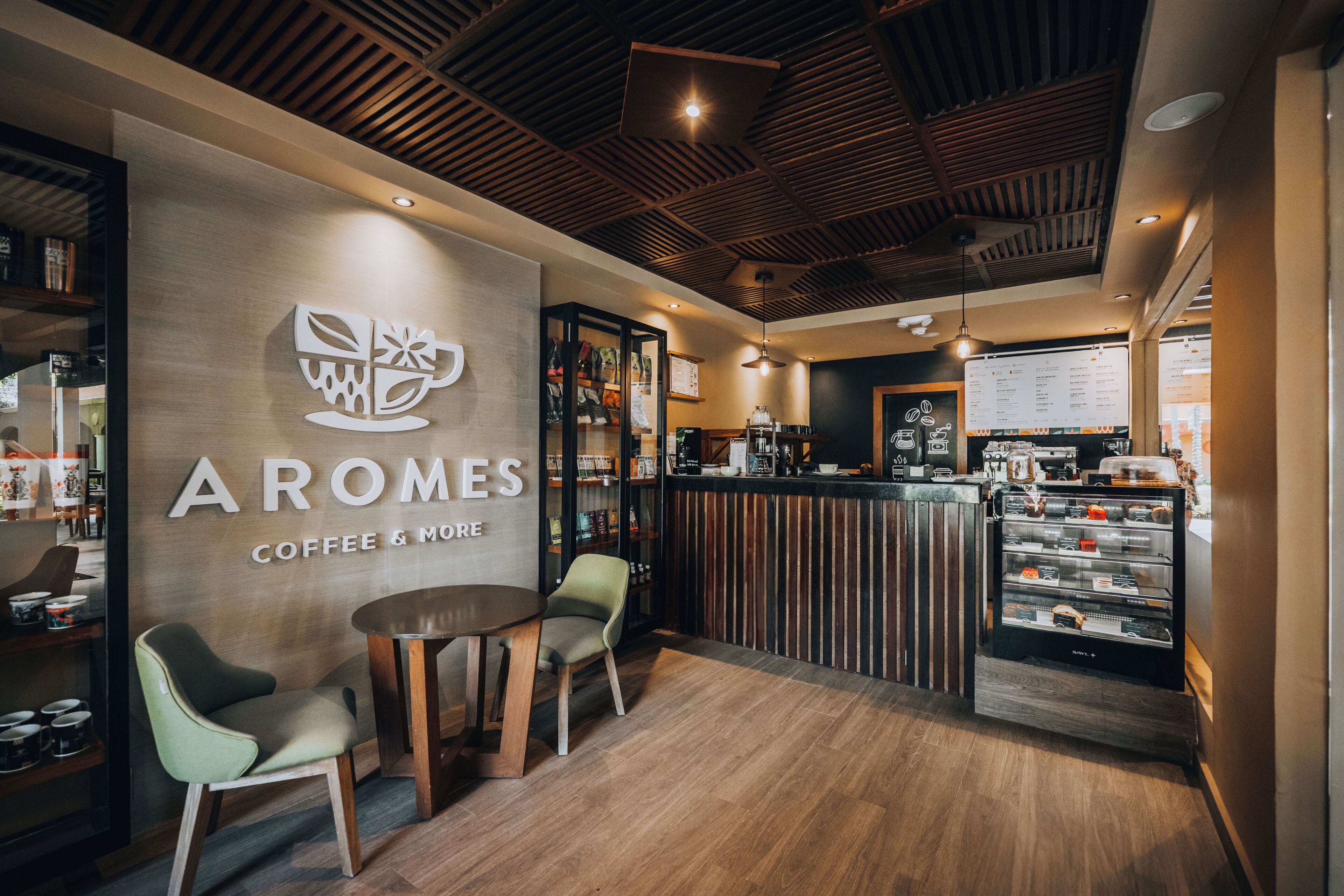 L’intérieur d’un café, avec un plafond en bois, un comptoir où sont exposées des pâtisseries et un espace salon.