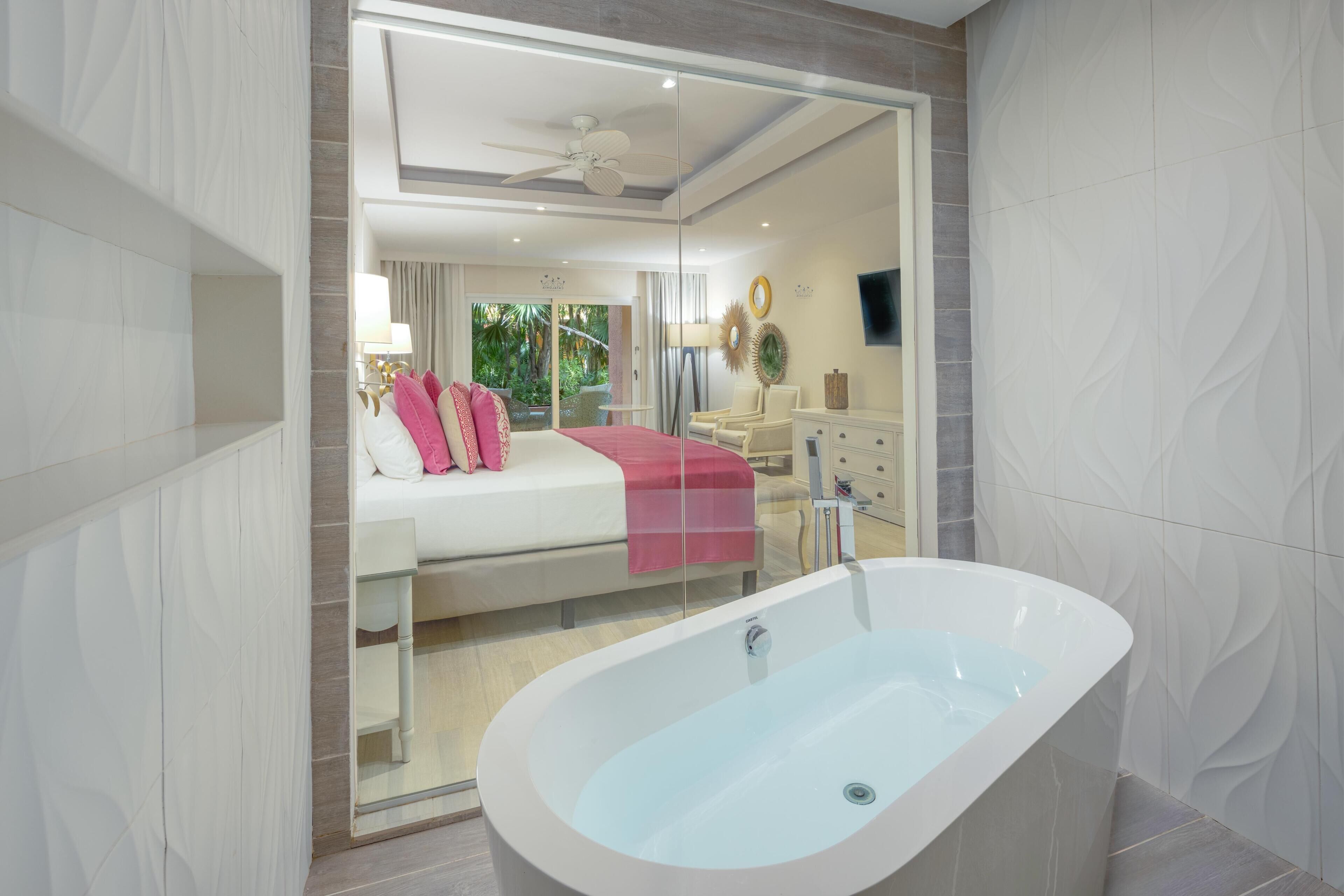 Un baño moderno con una bañera independiente, cabina de ducha con paredes de vidrio y una habitación con cama y manta roja a la vista.