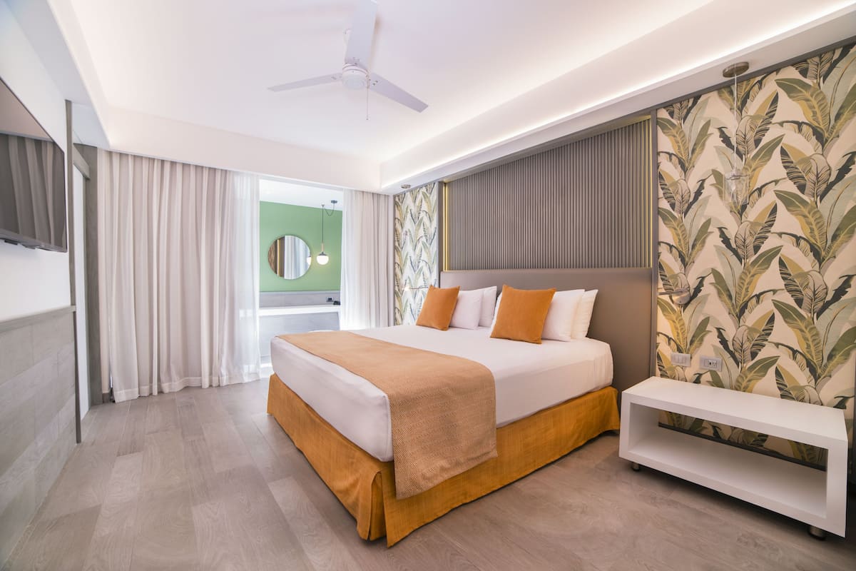 A modern hotel room with a large bed, a bedside table, a ceiling fan, and a view of the outdoors.