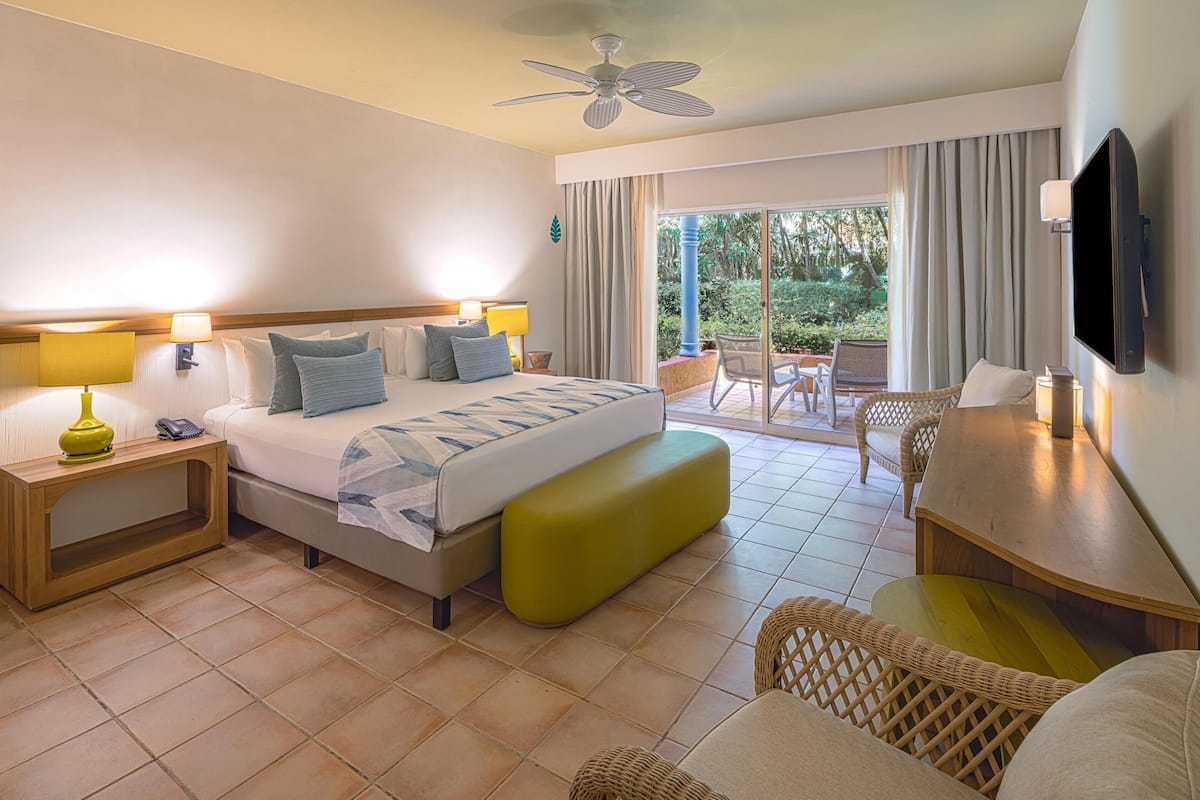 A hotel room with a bed, bedside tables, a TV, a balcony with chairs, and a ceiling fan.