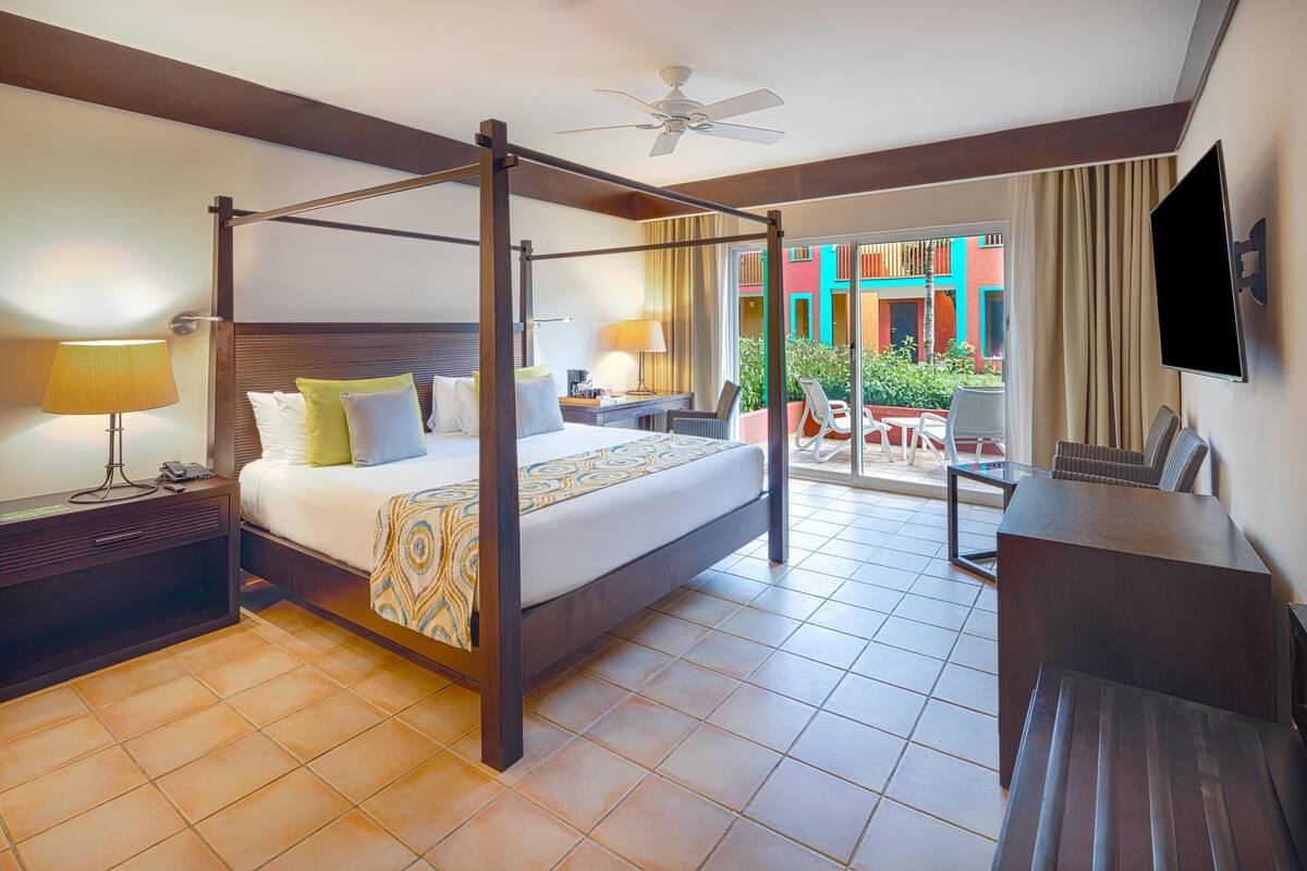 A four-poster bed with a desk and chair, a television, and a balcony with a view of colorful buildings.
