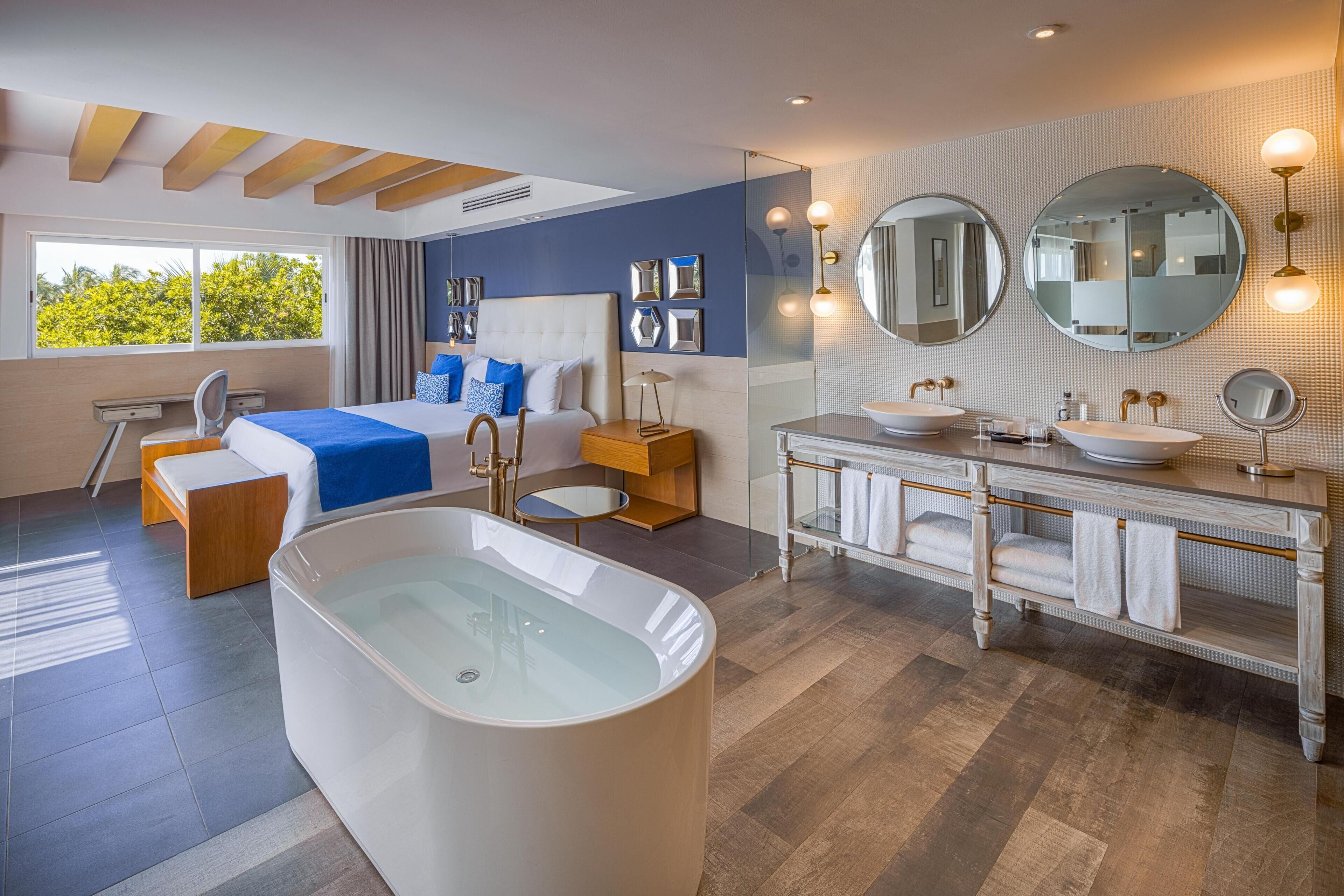 Une chambre d’hôtel moderne dotée d’une grande baignoire, d’un lit double, d’un bureau et d’une salle de bain avec un meuble-lavabo et deux vasques.