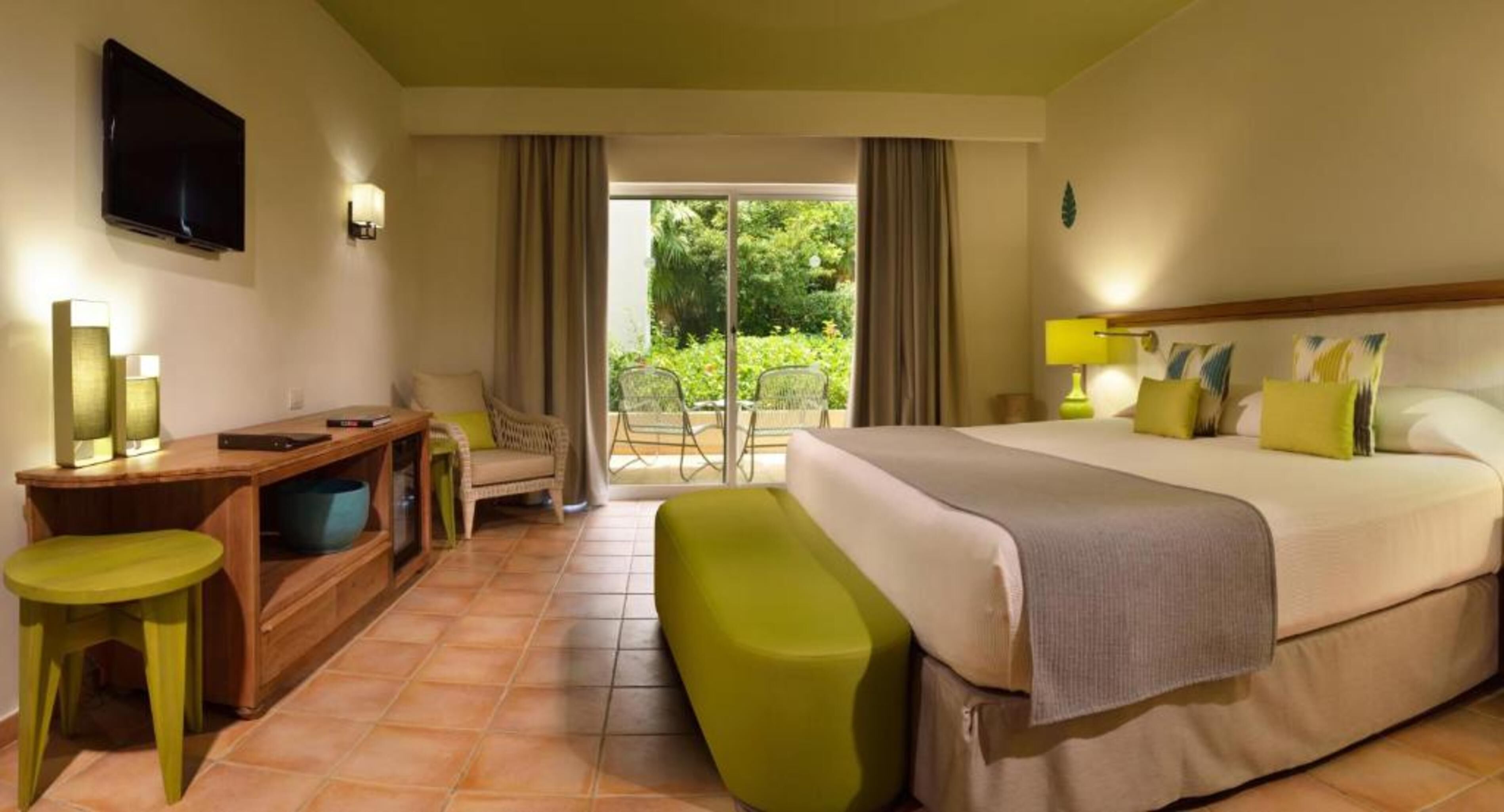 Ein Hotelzimmer mit Bett, Schreibtisch, Stuhl, Bank, Fernseher und Blick ins Freie.