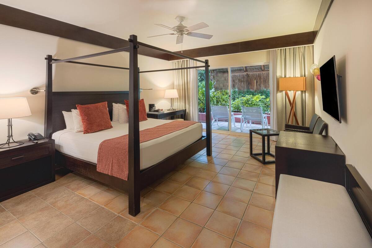A four-poster bed with a nightstand, a desk with a chair, a television, and a balcony with a chair and table.