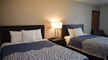 A hotel room with two beds, each with a patterned bedspread, a bedside table with a lamp, and a door in the background.