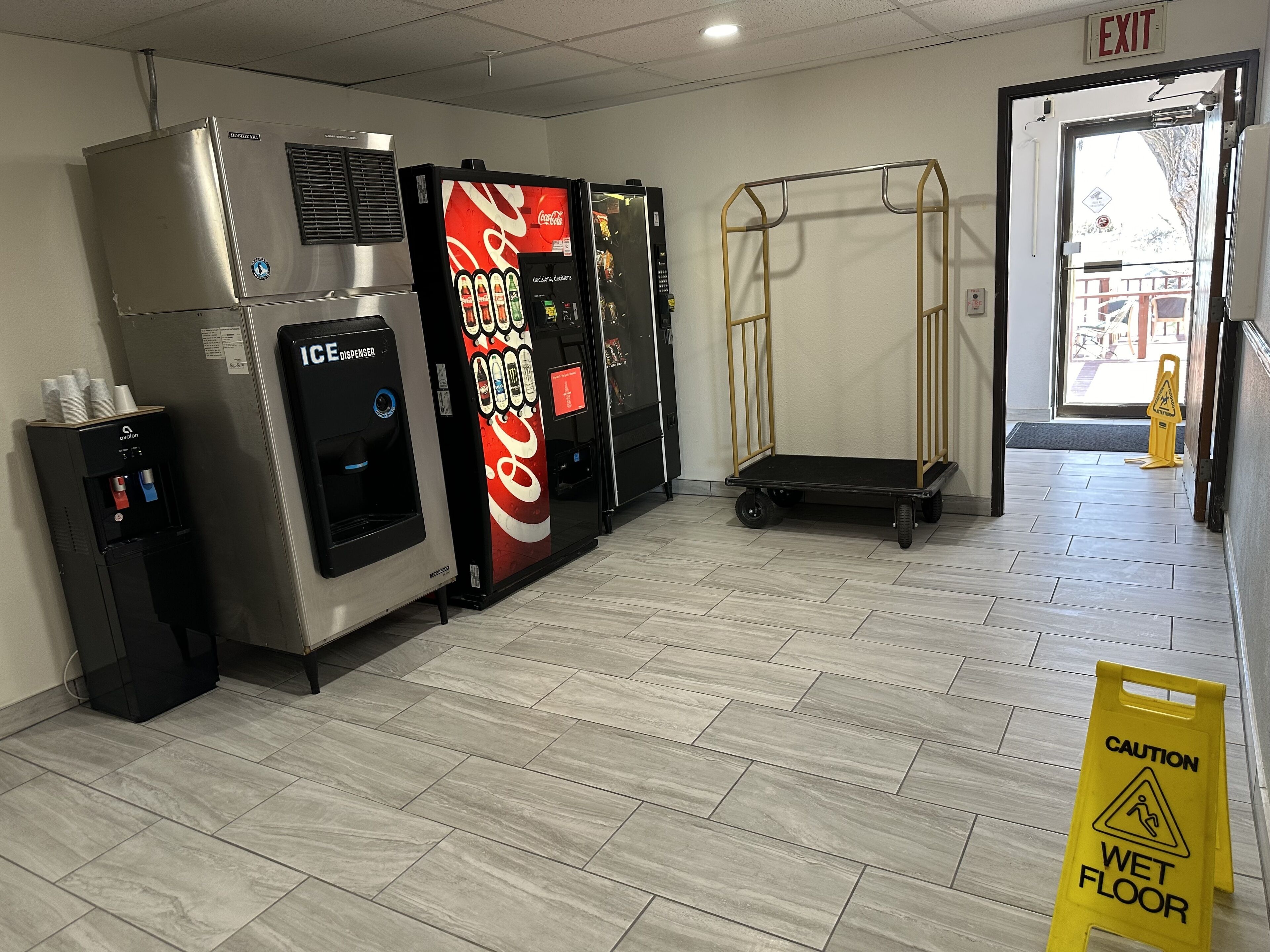 Un hall d’entrée propre et moderne, équipé d’un distributeur de Coca-Cola, d’un distributeur de glaçons et d’un distributeur d’eau. Un panneau d’avertissement indiquant un sol glissant est bien visible.