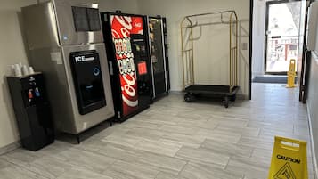 Un hall d’entrée propre et moderne, équipé d’un distributeur de Coca-Cola, d’un distributeur de glaçons et d’un distributeur d’eau. Un panneau d’avertissement indiquant un sol glissant est bien visible.
