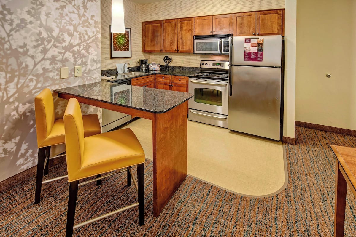 A kitchen with wooden cabinets, a stainless steel refrigerator, a microwave, and a stove. There is a small island with a glass top and two yellow chairs. The floor has a patterned carpet.