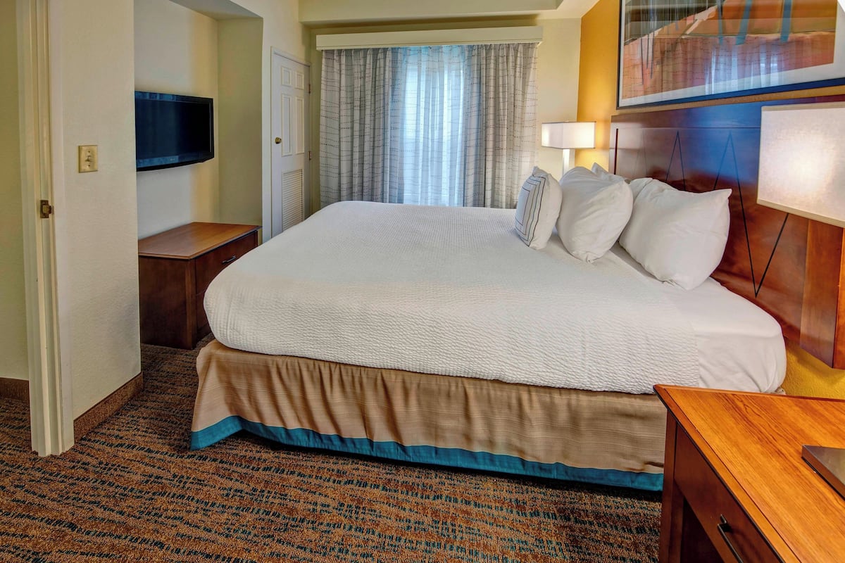 A hotel room with a large bed, a television mounted on the wall, a window with curtains, and a bedside table.