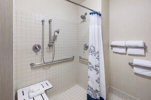 A small, tiled shower area with a grab bar, a showerhead, and a white shower curtain with a blue pattern. There are two white towels hanging on a towel rack.