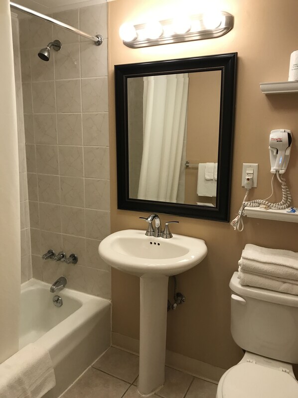 Un baño con bañera, lavamanos, inodoro, espejo y secador de pelo en un estante de pared.