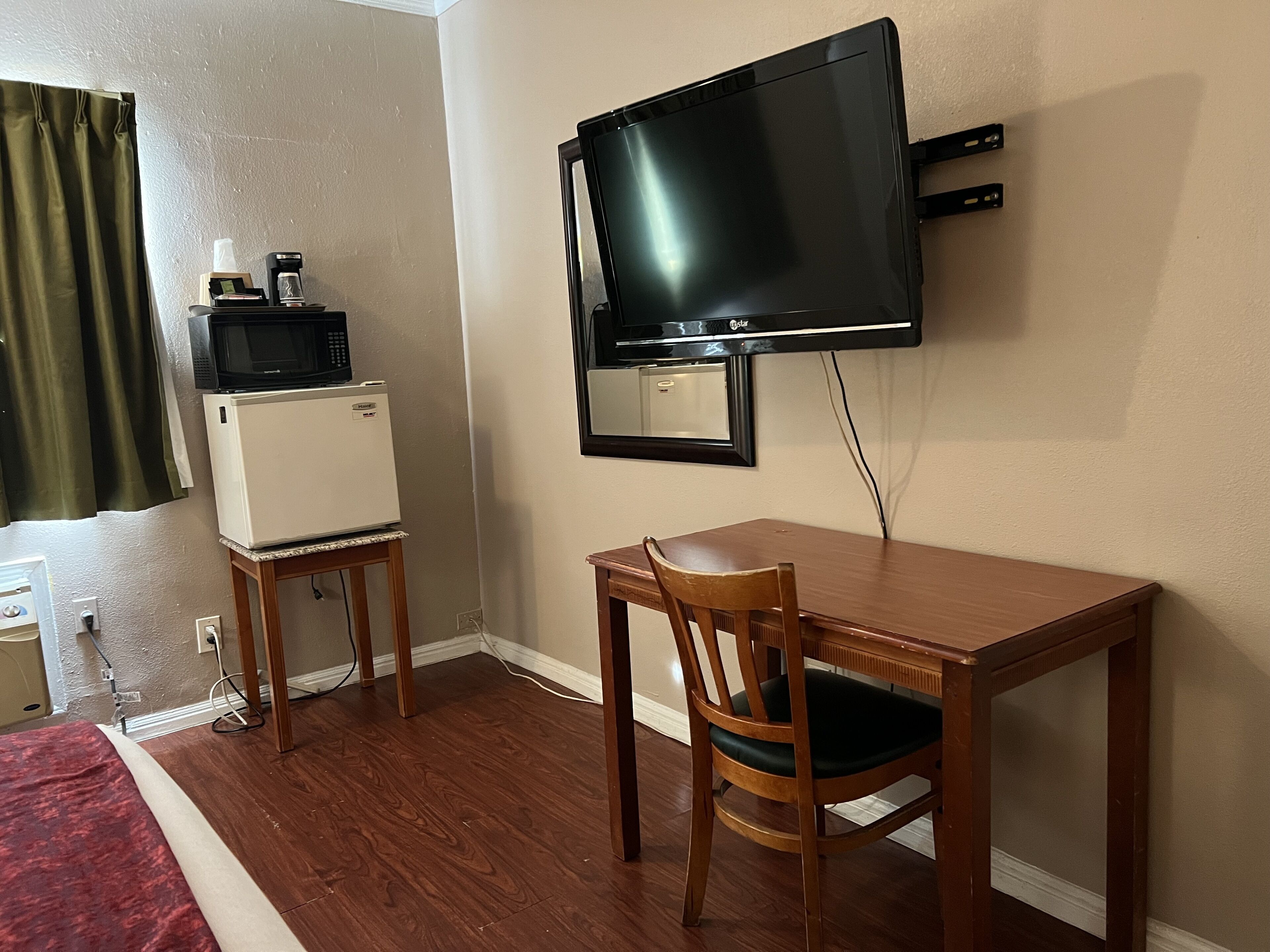 Una habitación pequeña con una mesa de madera, una silla, un televisor montado en la pared, un refrigerador y un microondas.