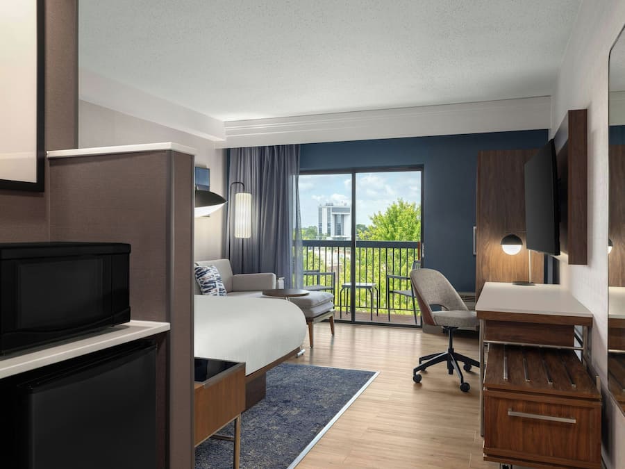 Una habitación de hotel moderna con cama, escritorio, silla, televisor y balcón con vistas a edificios y árboles.