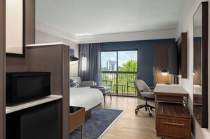 Una habitación de hotel moderna con cama, escritorio, silla, televisor y balcón con vistas a edificios y árboles.