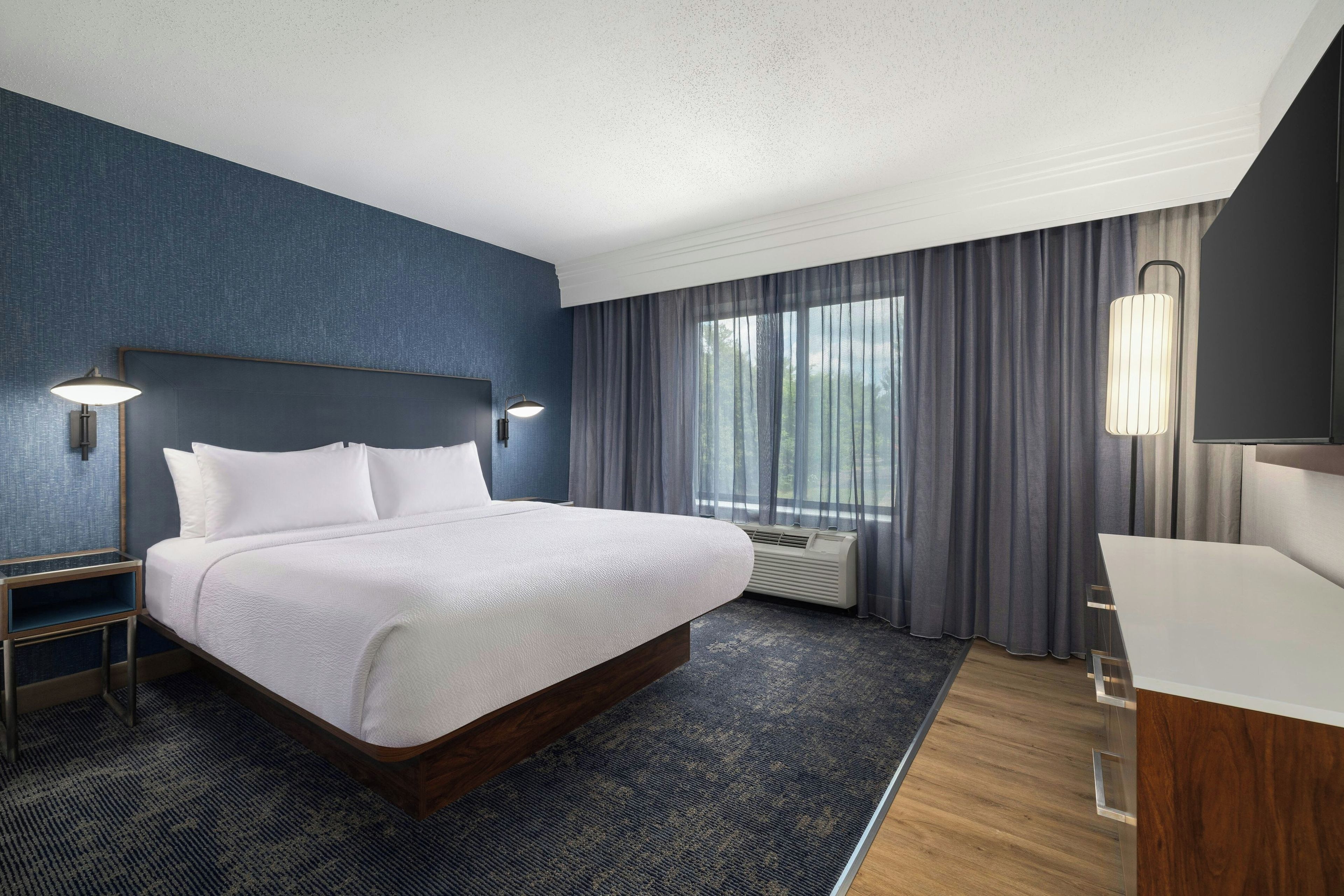 Habitación de hotel con una cama grande, una mesita de noche, un escritorio, una silla, un espejo y una ventana con cortinas.