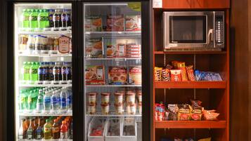 Máquina expendedora con bebidas y aperitivos, un microondas y un refrigerador con estantes llenos de alimentos.