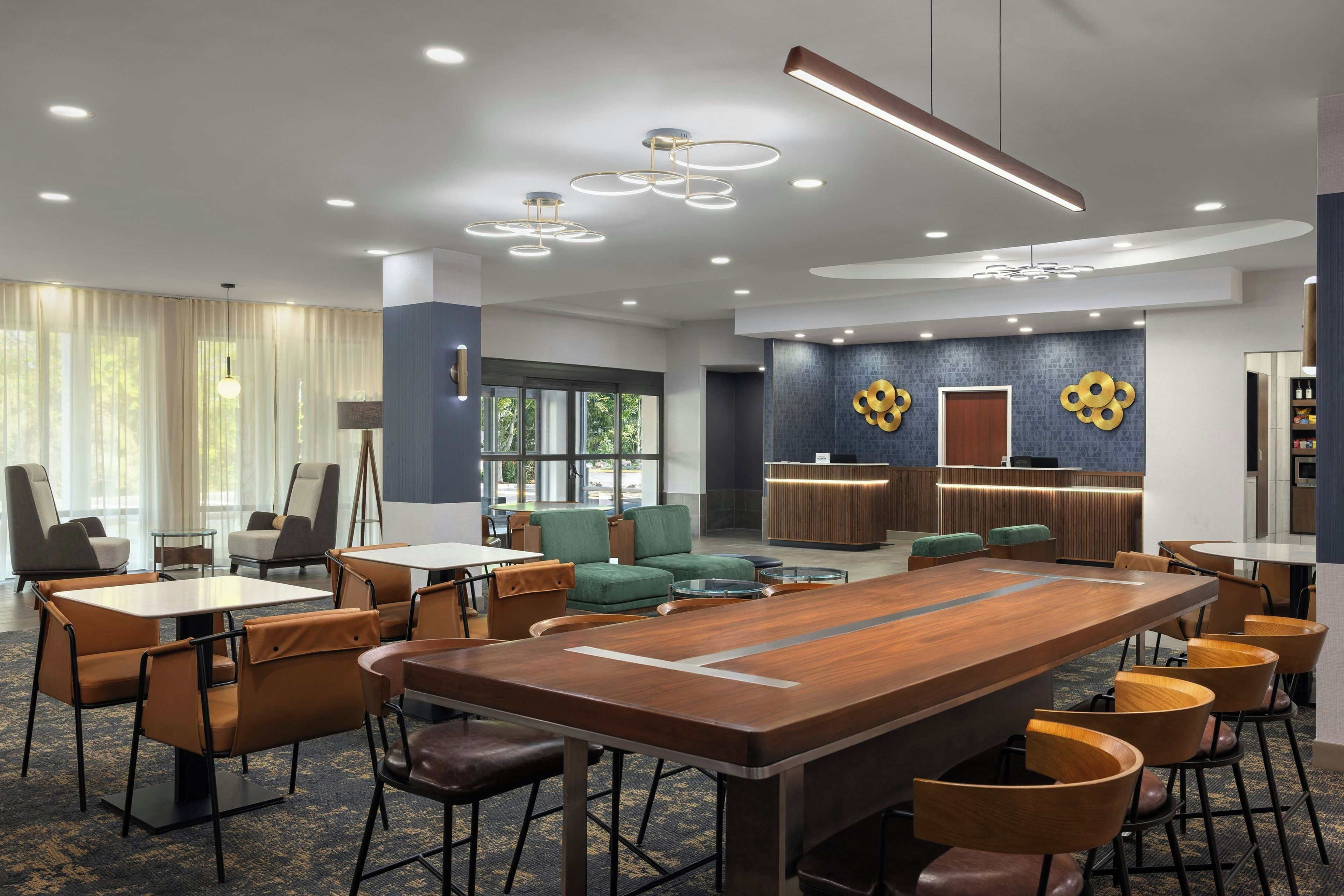 A modern hotel lobby with a central wooden conference table, seating arrangements, and a reception desk.