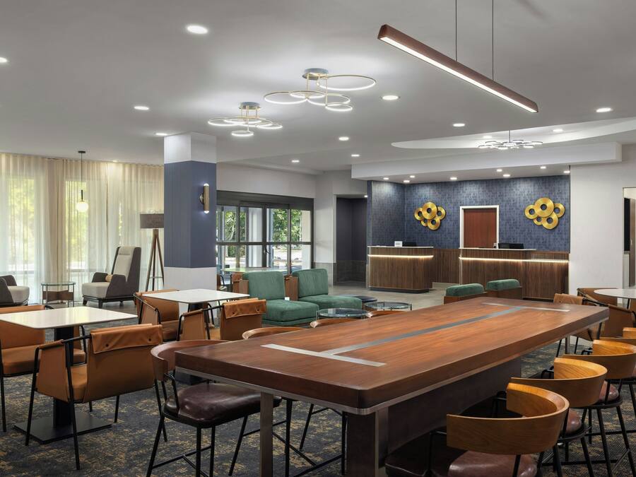 Um saguão de hotel moderno com uma mesa de conferência de madeira central, disposição de assentos e uma mesa de recepção.