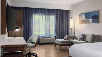 Habitación de hotel con cama, escritorio, silla, televisor, lámpara y ventana con cortinas.