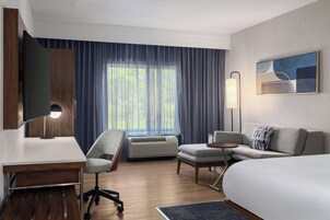 Habitación de hotel con cama, escritorio, silla, televisor, lámpara y ventana con cortinas.