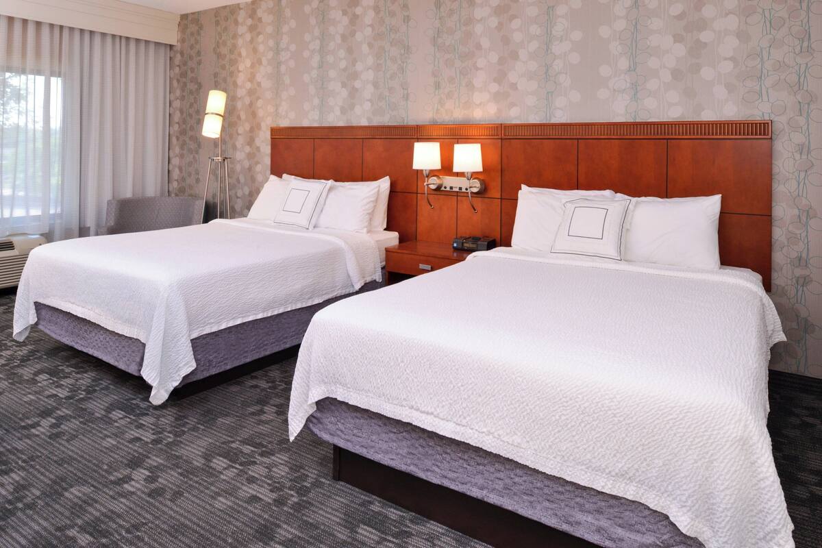 A hotel room with two beds, a nightstand, a lamp, and a chair.