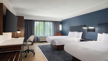 Une chambre d’hôtel avec deux lits, un bureau, une chaise, une lampe de bureau, un téléviseur fixé au mur et une fenêtre avec des rideaux.