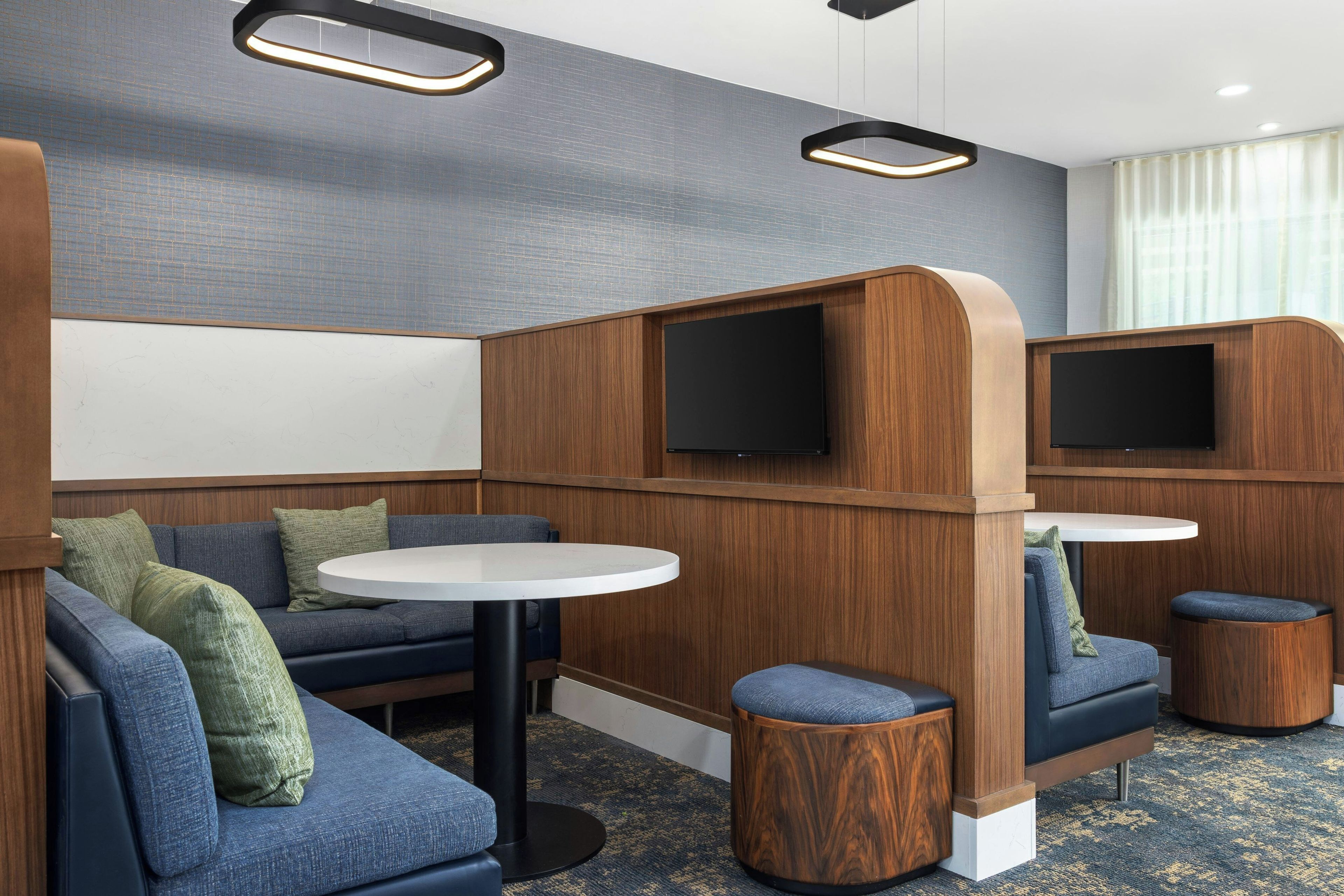 A modern hotel room with a sectional sofa, a round table, and two TV screens.