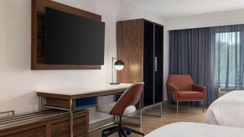 A hotel room with a large flat-screen TV mounted on the wall, a desk with a chair, a wardrobe, a single bed, and a window with curtains.