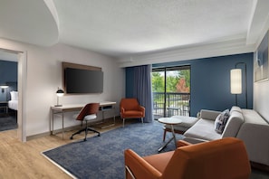 Quarto de hotel com escrivaninha, duas cadeiras, sofá, televisão e varanda com vista para árvores.