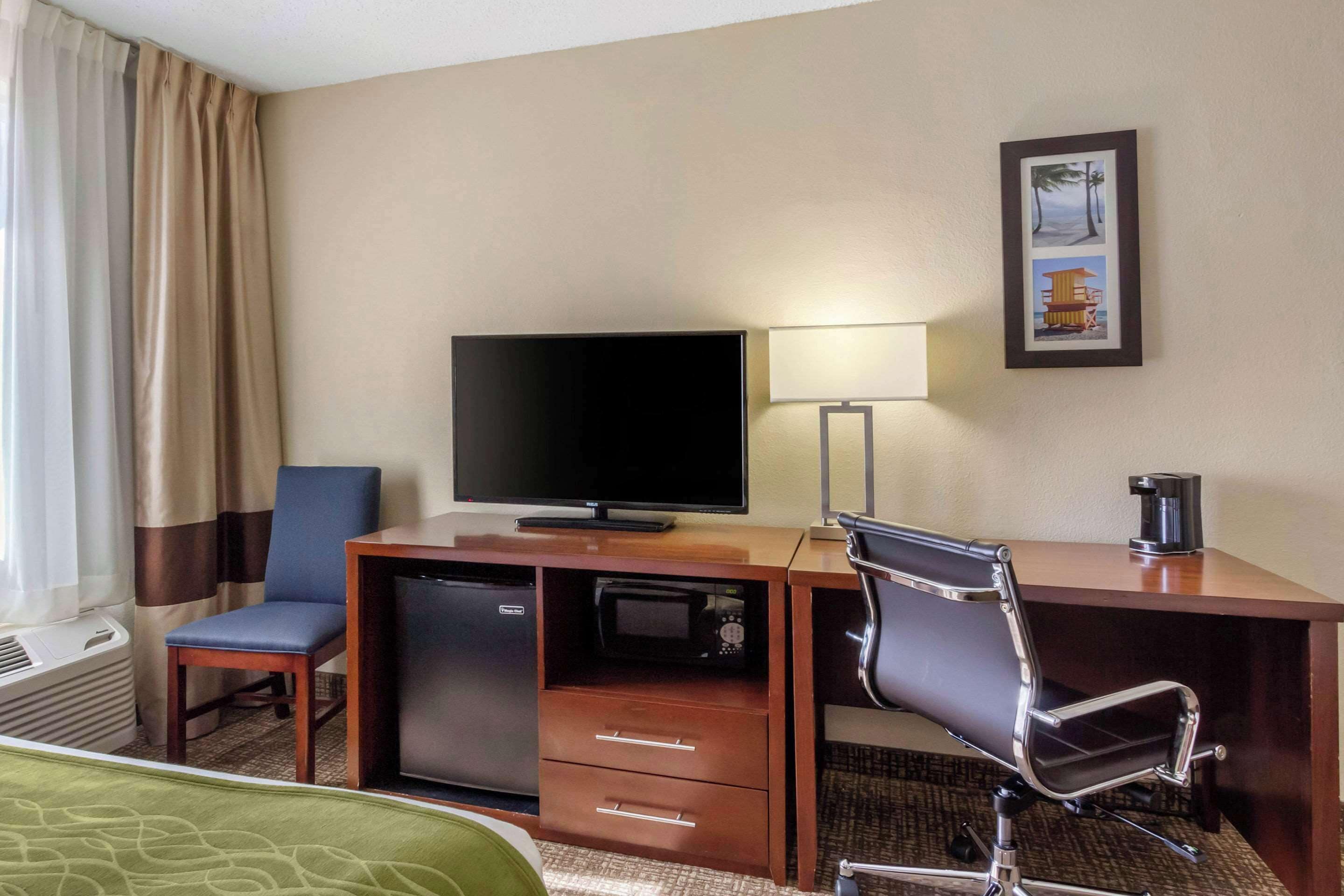 Habitación de hotel con cama, escritorio con televisor, silla, lámpara de escritorio, cafetera y un cuadro en la pared.