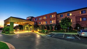 Ingresso di un hotel con area coperta, parcheggio e un edificio con numerose finestre.