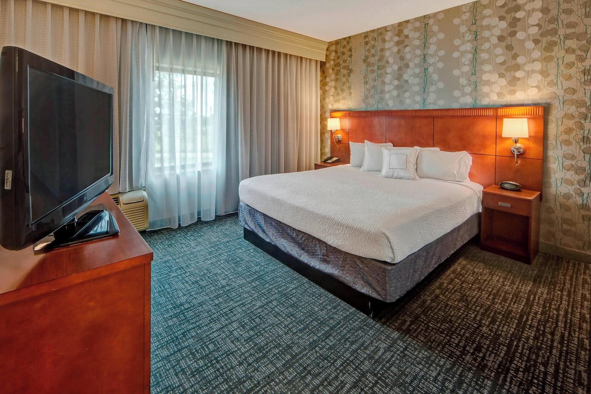 Une chambre d’hôtel avec un grand lit, un téléviseur posé sur une commode en bois et une table de chevet avec une lampe.