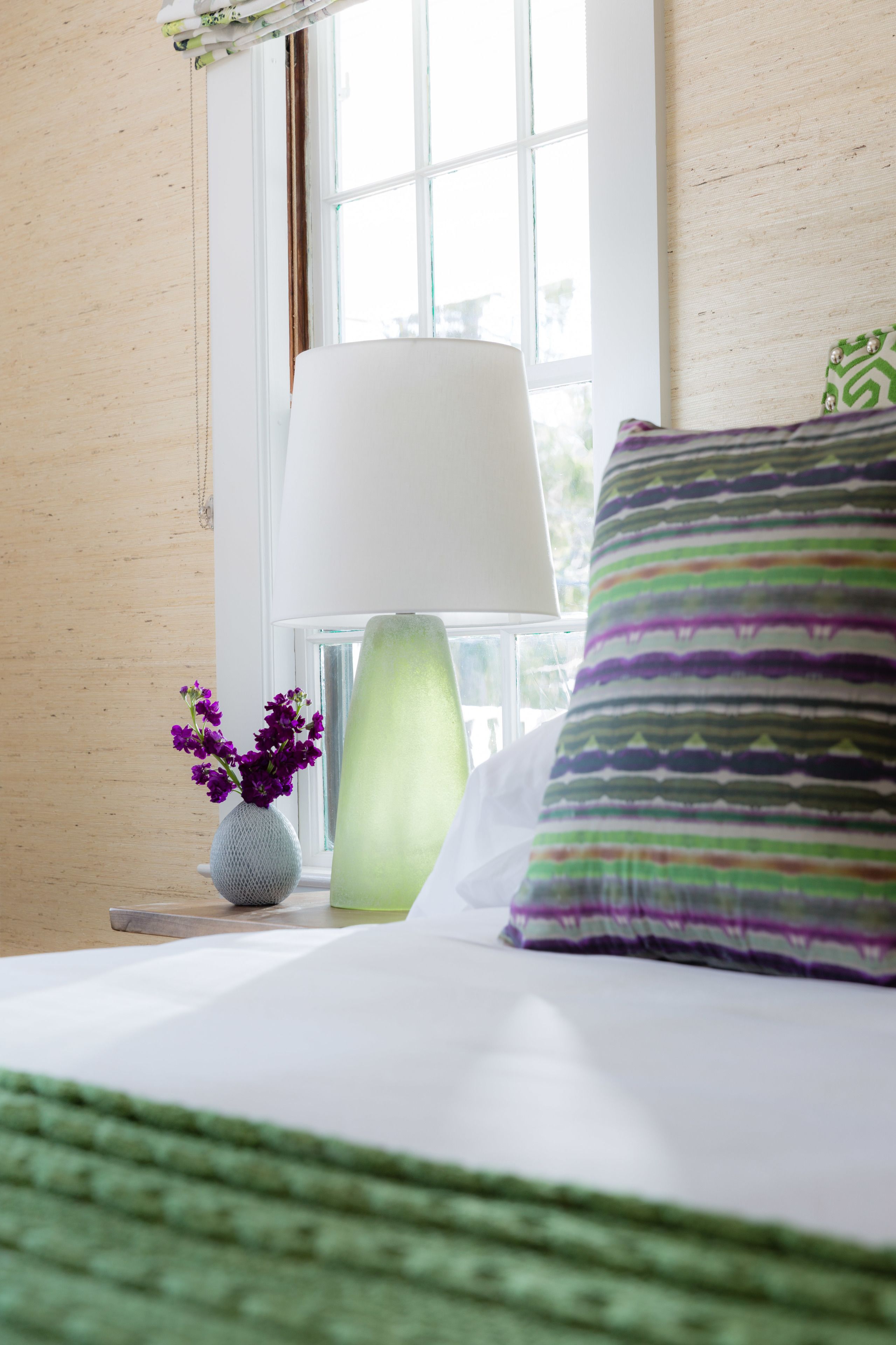A neatly made bed with a green blanket, a white pillow, and a striped pillow. A vase with purple flowers is on the nightstand beside the bed.